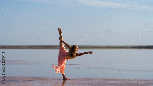 Flexible lady gymnast on the shore of a pink lake performs a split in a pink dress
