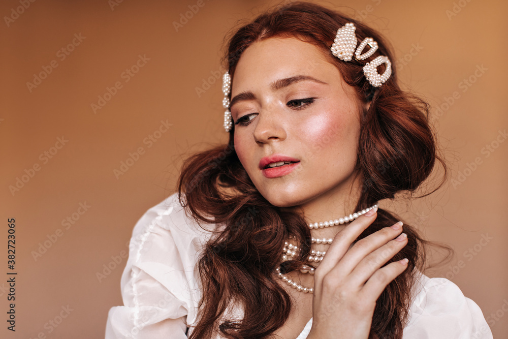 Lady with white hairpins in curly hair plays with her pearl necklace and gently looks down
