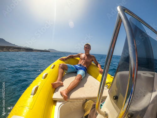 close up and portrait of young and happy beautiful man enjoying summer lying down on a small boat or dinghy in the middle of the sea - male with abs smiling looking at the camera in his vacations