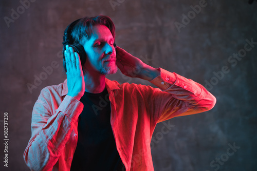 Adult hipster man with long hair in big headphones and shirt with red and blue neon lighting on dark background. Nice music concept.