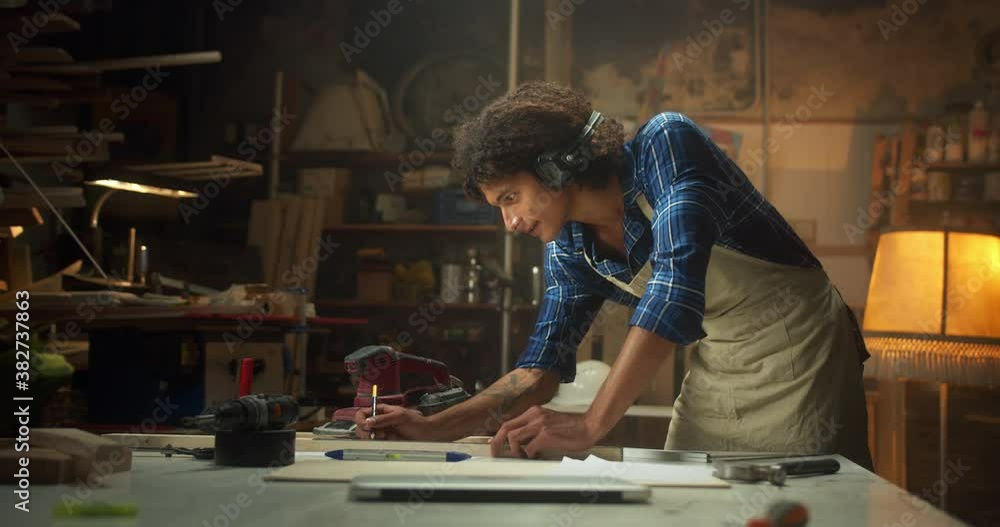 Side shot of pleasant carpenter drawing design carpentry project at cozy workshop indoors, attractive curly young man in apron and caged shirt leaning on work desk instruments engineer concept Side shot of pleasant carpenter drawing design carpentry project at cozy workshop indoors, attractive curly young man in apron and caged shirt leaning on work desk instruments engineer concept