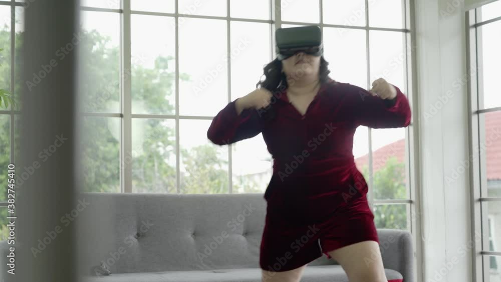 Asian plus size woman in VR glasses with joystick playing virtual game in living room at home Asian plus size woman in VR glasses with joystick playing virtual game in living room at home