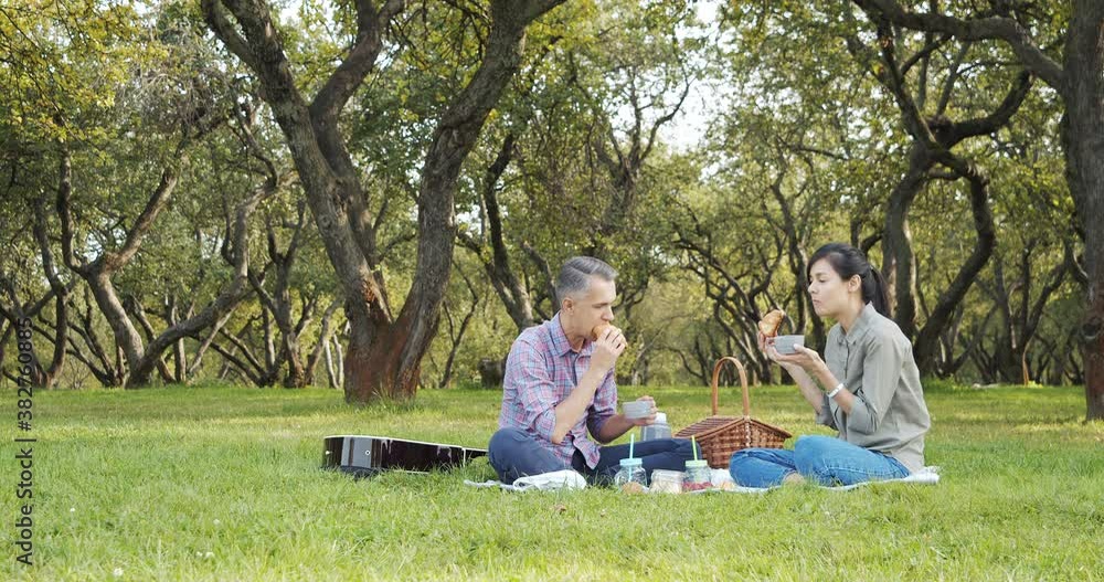 Happy couple spending time together drinking hot tea from a thermos and eating croissants on a picnic in a green garden. Happy couple spending time together drinking hot tea from a thermos and eating croissants on a picnic in a green garden.
