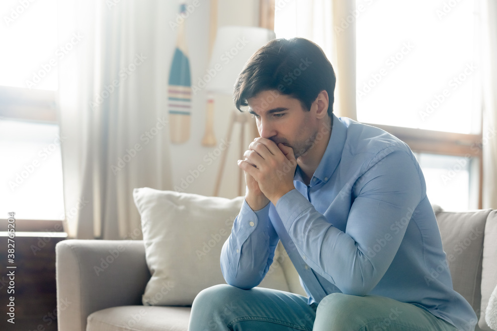 What should I do. Pensive sad millennial guy sitting alone on sofa in living room leaning forward clasping hands at chin lost in serious unhappy thoughts, looking for way out of difficult situation What should I do. Pensive sad millennial guy sitting alone on sofa in living room leaning forward clasping hands at chin lost in serious unhappy thoughts, looking for way out of difficult situation