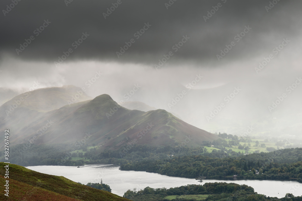 Stunning epic landscape image across Derwentwater valley with falling rain drifting across the mountains causing pokcets of light and dark across the countryside