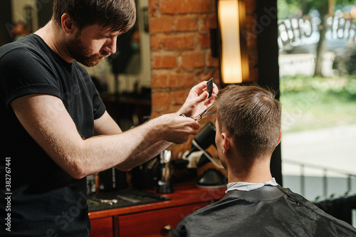 Bearded hairdresser making a haircut for a young man in a barber shop