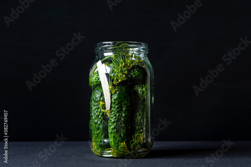 Pickled cucumbers with herbs. Marinated cucumber gherkins in glass jar on black background with copy space. Homemade preserves