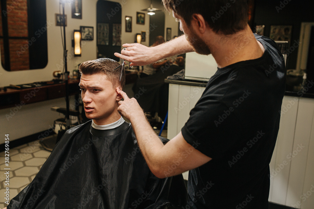 Hairdresser folding ear, making a haircut for a young man in a barber