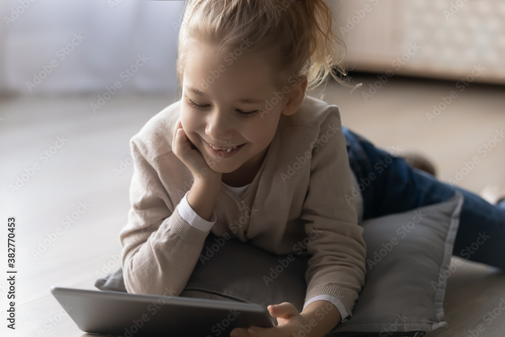 Close up smiling little girl using tablet, lying on warm wooden floor with underfloor heating at home, happy pretty child holding gadget, browsing apps, playing game, watching funny video Close up smiling little girl using tablet, lying on warm wooden floor with underfloor heating at home, happy pretty child holding gadget, browsing apps, playing game, watching funny video