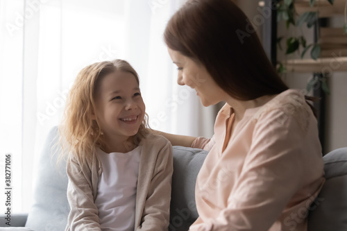 Overjoyed adorable little girl with loving young mother having fun at home, sitting on couch, caring mum hugging excited pretty daughter, chatting, talking, sharing secrets, spending leisure time