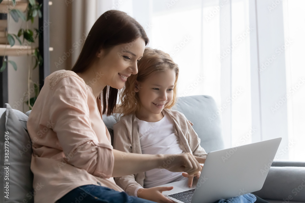 Smiling young mother teaching little daughter to use laptop, pointing finger at screen, sitting on cozy couch at home, happy mum and adorable pretty girl having fun, watching video, browsing apps Smiling young mother teaching little daughter to use laptop, pointing finger at screen, sitting on cozy couch at home, happy mum and adorable pretty girl having fun, watching video, browsing apps