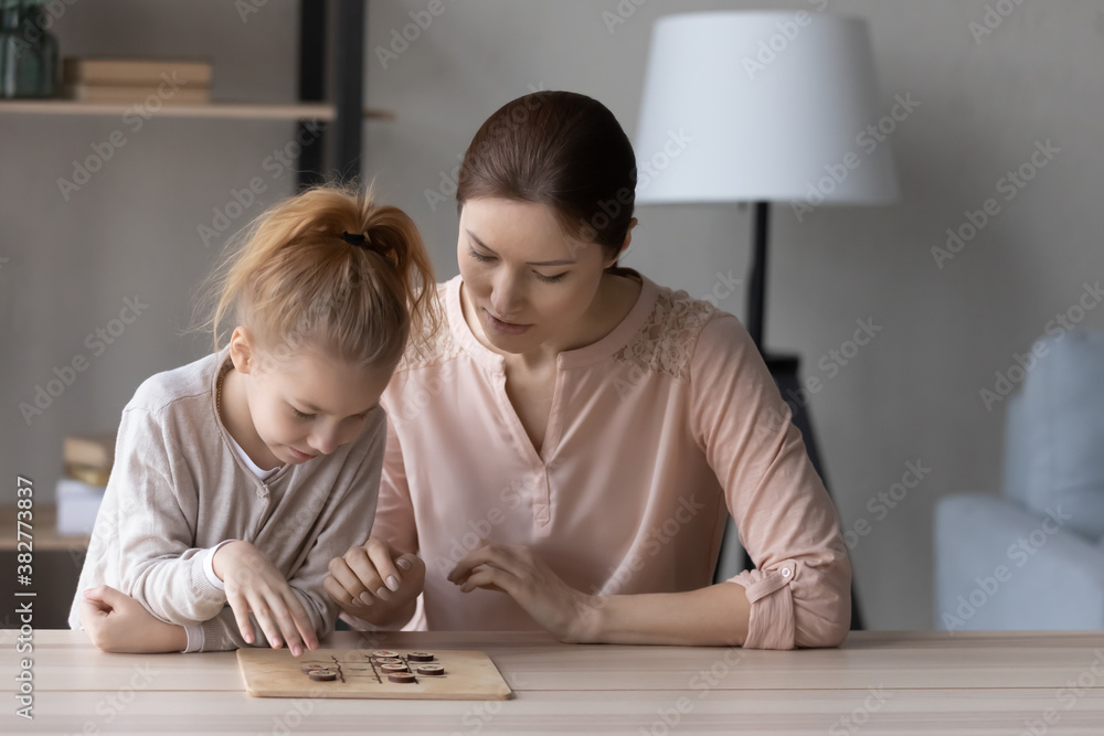 Pretty little girl and mother playing interesting wooden board game, checkers on weekend, sitting at table at home, focused young woman and preschool daughter spending leisure time together Pretty little girl and mother playing interesting wooden board game, checkers on weekend, sitting at table at home, focused young woman and preschool daughter spending leisure time together