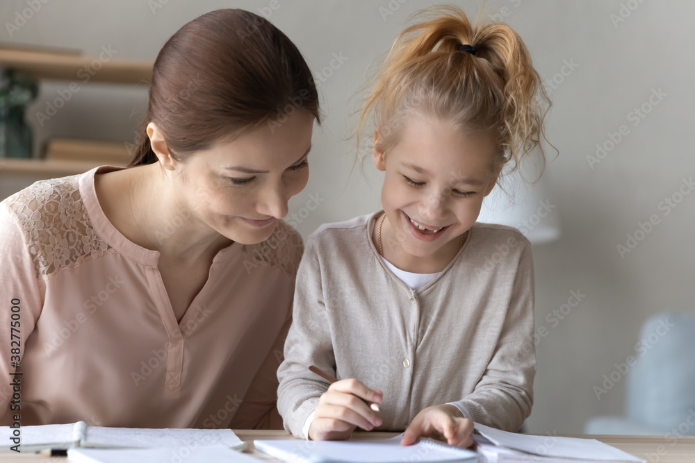 Caring mother and smiling little girl doing school homework together, sitting at desk at home, young mum and daughter studying, writing in notebook, teacher tutor babysitter teaching pupil Caring mother and smiling little girl doing school homework together, sitting at desk at home, young mum and daughter studying, writing in notebook, teacher tutor babysitter teaching pupil