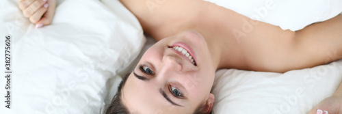 Top view of smiling brunette lady laying naked under blanket. Clean white bedding. Happy young woman wake up. Upside down female. Morning routine and relaxation concept photo