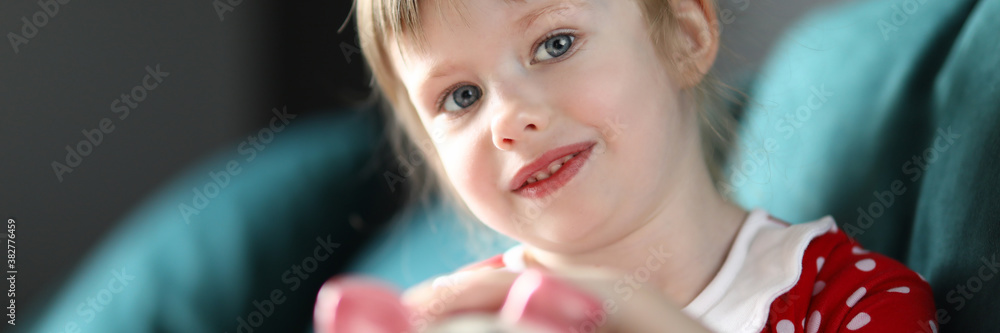 Portrait of smiling beautiful child holding piggy bank sitting on sofa at home. Lovely blonde kid saving up for future. Girl in red dress. Childhood and happiness concept Portrait of smiling beautiful child holding piggy bank sitting on sofa at home. Lovely blonde kid saving up for future. Girl in red dress. Childhood and happiness concept