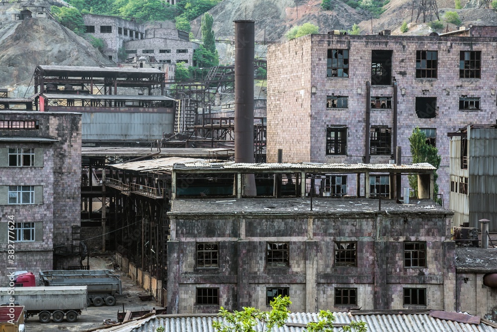 View on abandoned factory in the mountains covered with green trees in Armenia. Many brick buildings with windows, a pipe and cargo cars. View on abandoned factory in the mountains covered with green trees in Armenia. Many brick buildings with windows, a pipe and cargo cars.
