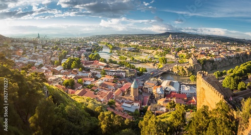 Aerial view from the Narikala fortress on the Tbilisi city, the capital of Georgia, full of small tiled roof houses, green trees, bridges over the Kura river in a sunny day.