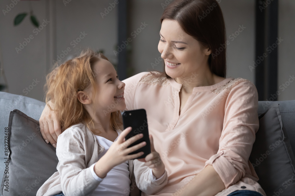 Smiling cute little girl and young mother having fun with phone, sitting on cozy couch at home, happy family enjoying leisure time together, shopping online, watching video, browsing apps Smiling cute little girl and young mother having fun with phone, sitting on cozy couch at home, happy family enjoying leisure time together, shopping online, watching video, browsing apps