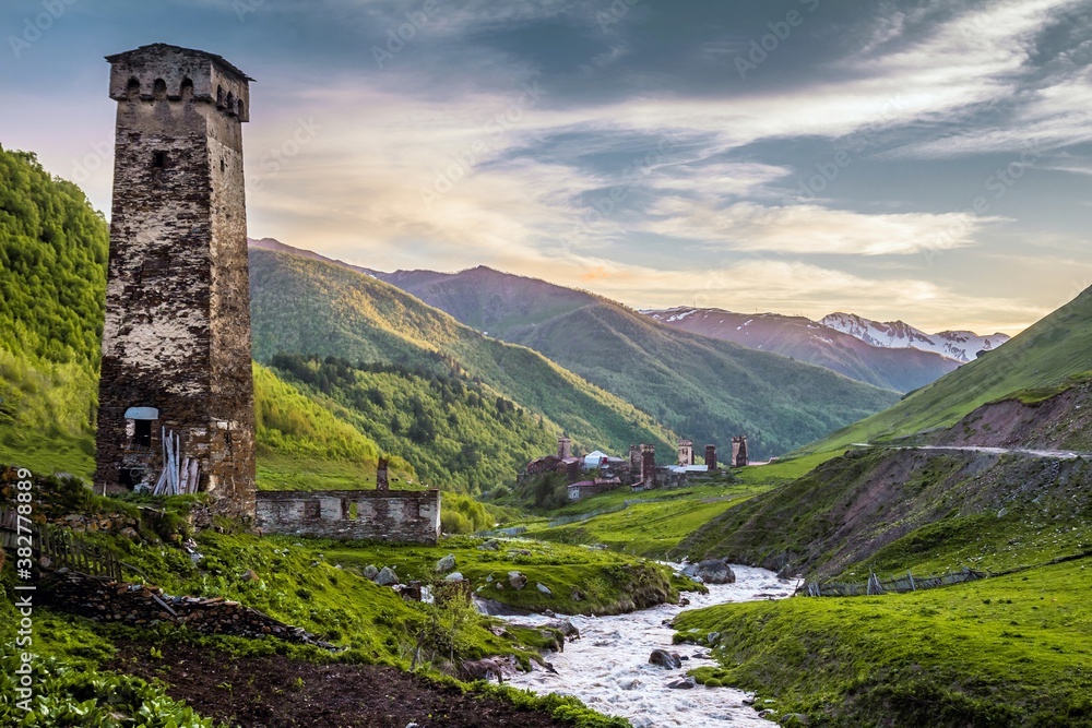 Obraz premium Gorgeous landscape in the Ushguli village, Georgia: medieval tower in the downhill on the bank of a rapid narrow river and green hills against pink sky in the background.