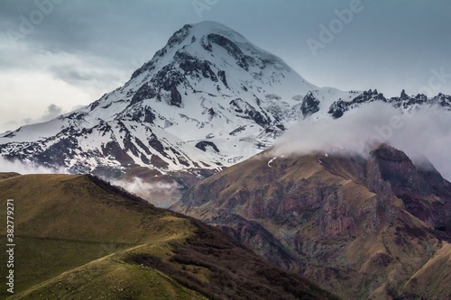 Gorgeous view on Caucasus Kazbek mount located on the border of Georgia and Russia, covered with snow and small grassy hills in clouds in the foreground.