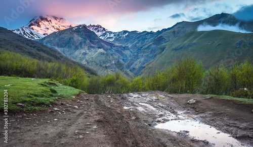 Gorgeous view on Caucasus Kazbek mount covered with snow at the sunrise, small hills in clouds and muddy road with car traces in the foreground, Georgia.