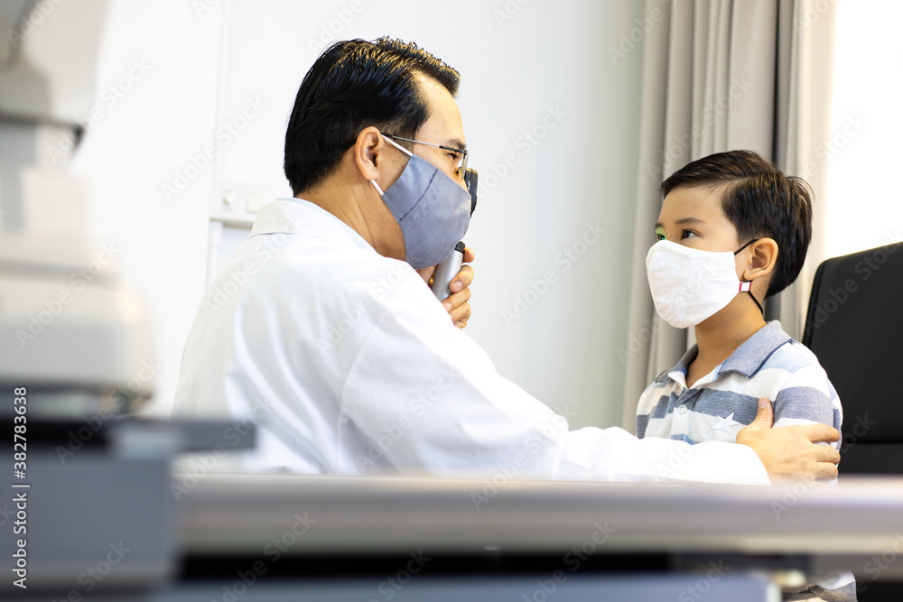 An Asian male doctor uses An ophthalmoscope to examine the boy's eyes. The ophthalmologist examines the child's vision. They wear a protective face mask. An Asian male doctor uses An ophthalmoscope to examine the boy's eyes. The ophthalmologist examines the child's vision. They wear a protective face mask.