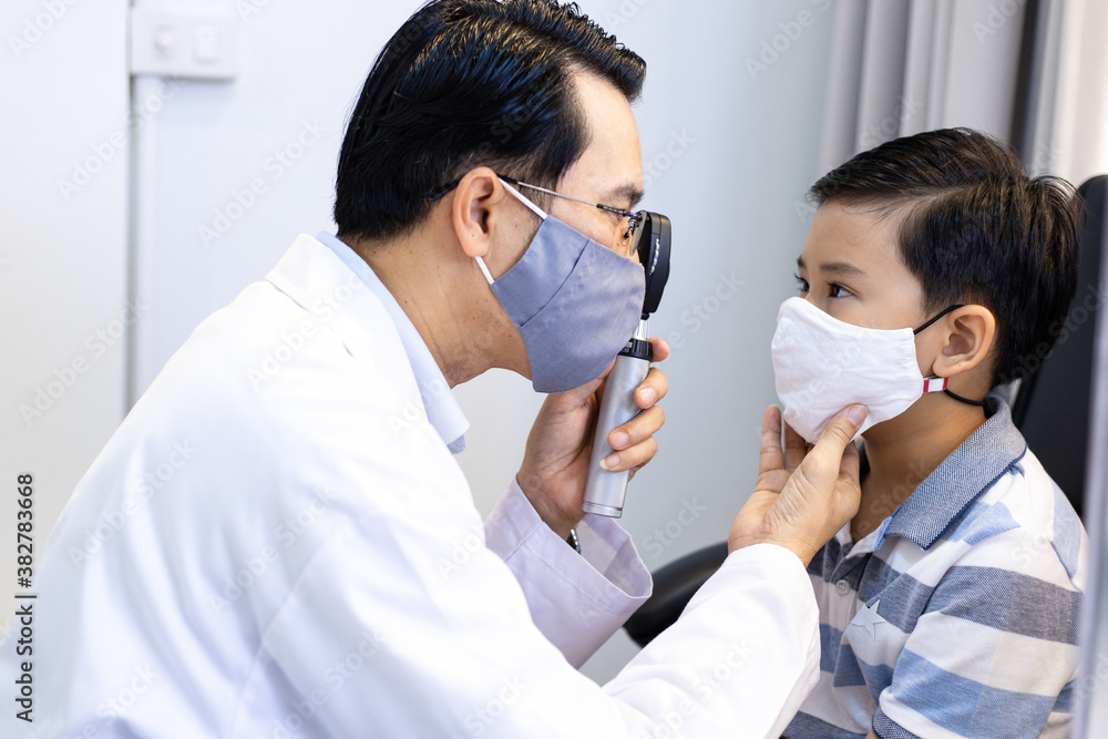 An Asian male doctor uses An ophthalmoscope to examine the boy's eyes. The ophthalmologist examines the child's vision. They wear a protective face mask.