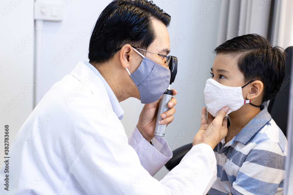 An Asian male doctor uses An ophthalmoscope to examine the boy's eyes. The ophthalmologist examines the child's vision. They wear a protective face mask. An Asian male doctor uses An ophthalmoscope to examine the boy's eyes. The ophthalmologist examines the child's vision. They wear a protective face mask.