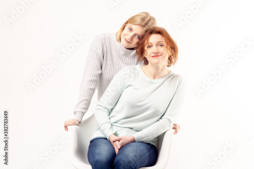 Young teenage blonde girl hugging her smiling ginger hair senior mother sitting on the chair