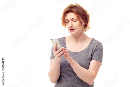 Red haired adult mature woman in gray dress looking on mobile phone in her hands with pensive expression
