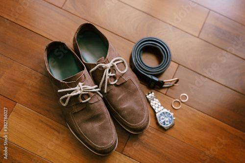 Wooden floor with brown men's shoes, wristwatch, belt and wedding rings.