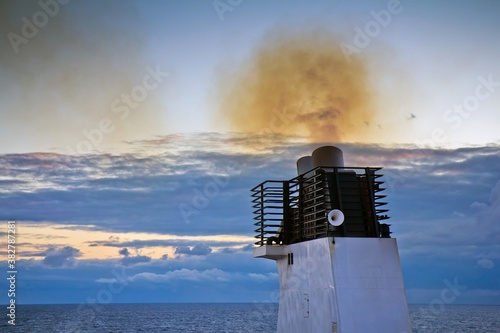 Closeup of funnel of a cargo ship floating in the Baltic sea with smoke polluting environment against cloudy sky in the evening.