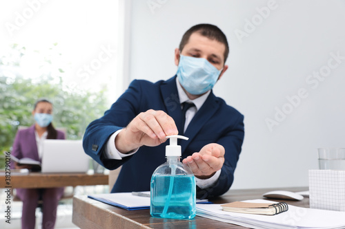 Office employee in mask applying hand sanitizer at workplace