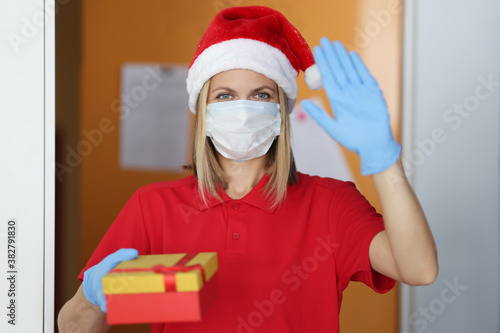 Woman postman in santa claus hat in medical mask and rubber gloves holds gift in her hand and waves hello. Contactless delivery during covid19 pandemic concept. © H_Ko