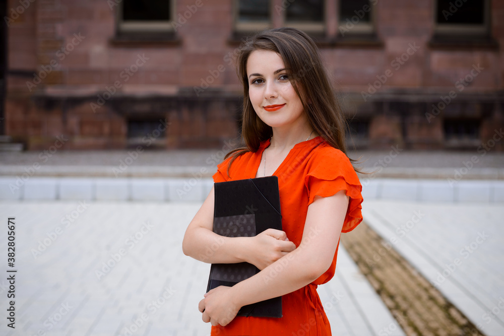 Beautiful confident young brunette woman in red dress stands on the street, holds folder in her hands, looks to the camera and smiles. Female student near the university. Beautiful confident young brunette woman in red dress stands on the street, holds folder in her hands, looks to the camera and smiles. Female student near the university.