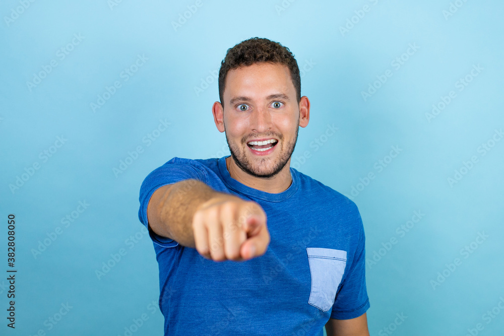 Young handsome man wearing blue casual t-shirt over isolated blue background laughing at you, pointing finger to the camera with hand over body, shame expression Young handsome man wearing blue casual t-shirt over isolated blue background laughing at you, pointing finger to the camera with hand over body, shame expression