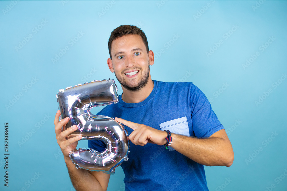 Fototapeta premium Young handsome man wearing blue casual t-shirt over isolated blue background holding a number five balloon. Celebration concept