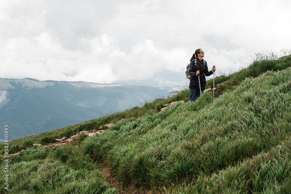 A young woman with hiking sticks climbs to the top of the mountain in a hiking wear and enjoying with amazing landscapes of a mountains around her