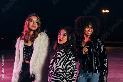 Fashionable girls standing together on roof at night under neon lights. Multi-ethnic group of women posing outdoors and looking at camera.