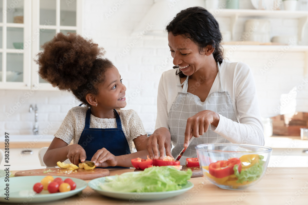 Funny little african ethnicity kid girl wearing apron, talking to affectionate caring biracial mommy while preparing healthy food together, enjoying improving culinary skills in modern kitchen.