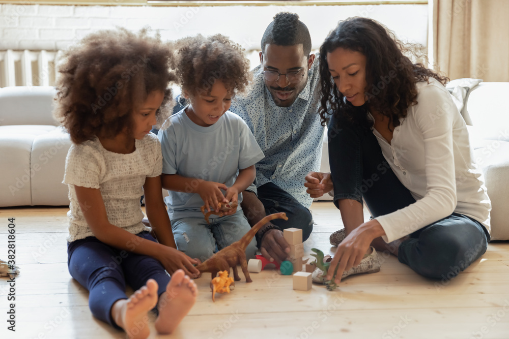 Full length happy african american married couple playing toys with cheerful small children siblings, sitting together on warm wooden floor in living room, affectionate loving family weekend pastime. Full length happy african american married couple playing toys with cheerful small children siblings, sitting together on warm wooden floor in living room, affectionate loving family weekend pastime.