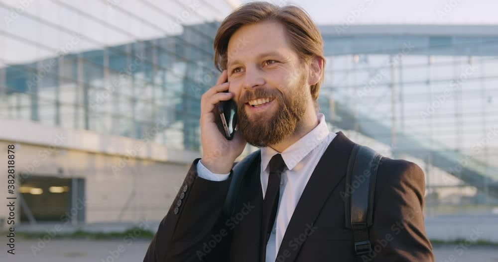 Portrait of good-natured smiling stylish successful young man with beard which talking on phone near big modern city glass construction Portrait of good-natured smiling stylish successful young man with beard which talking on phone near big modern city glass construction
