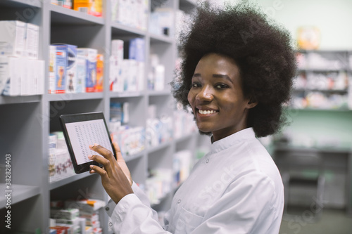 Young pretty smiling African American dark skinned female pharmacist using digital tablet during inventory, or making order of medicines in modern hospital pharmacy.