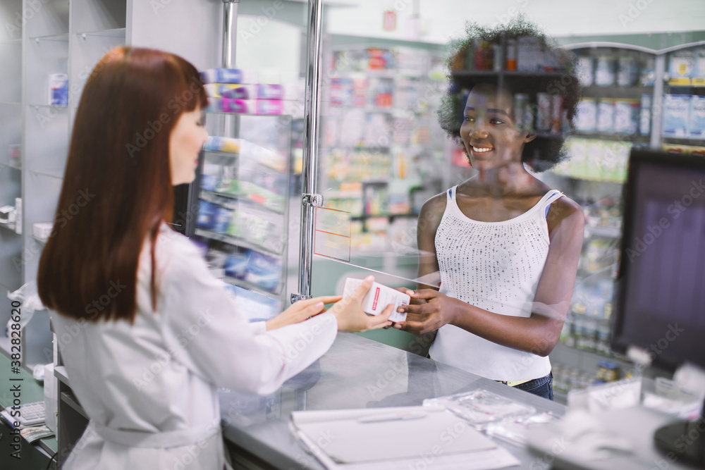 Back view of beautiful young Caucasian pharmacist selling medications to pretty smiling African lady patient. Medicine dispensing in modern pharmacy. Healthcare concept