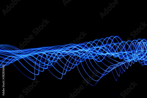 Long exposure photograph of neon electric blue colour in an abstract swirl, parallel lines pattern against a black background. Light painting photography.