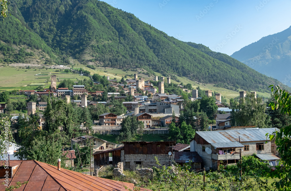 Panoramic view of Mestia, a highland townlet in Georgia in the Caucasus Mountains. 
Houses with stone defensive towers on the foreground and surrounding mountains with green forest on the background.