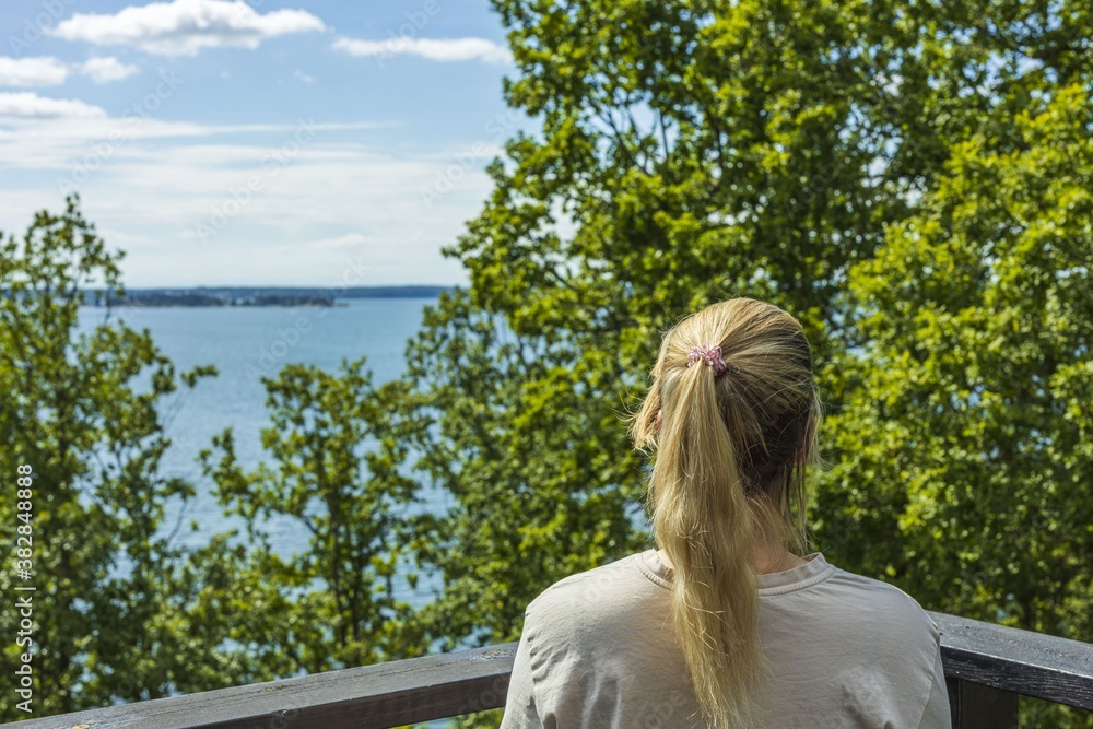 Beautiful summer Baltic sea landscape view. Young blond woman looking on sea through green tree tops. 