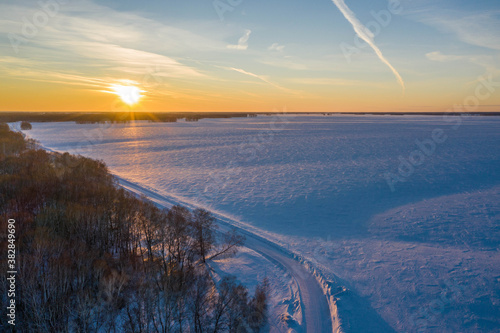 Aerial shot. A winter country road along a frozen field against the backdrop of a sunset