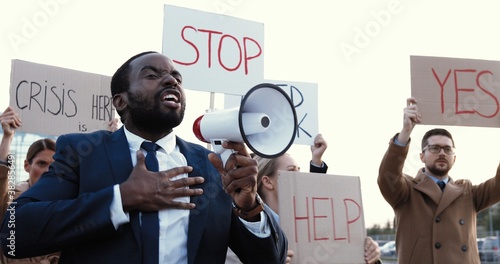 African American handsome man in suit and tie talking in megaphone outdoor at protest against unemployment in multi ethnic crowd. Male having speech at strike against inequality and for human rights. photo