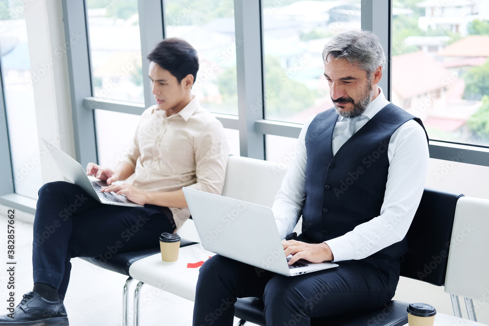 Successful Senior businessman is working with laptop in workspace at office. He is sitting distance from asian staff. Technology for business and New normal lifestyle concept. Successful Senior businessman is working with laptop in workspace at office. He is sitting distance from asian staff. Technology for business and New normal lifestyle concept.